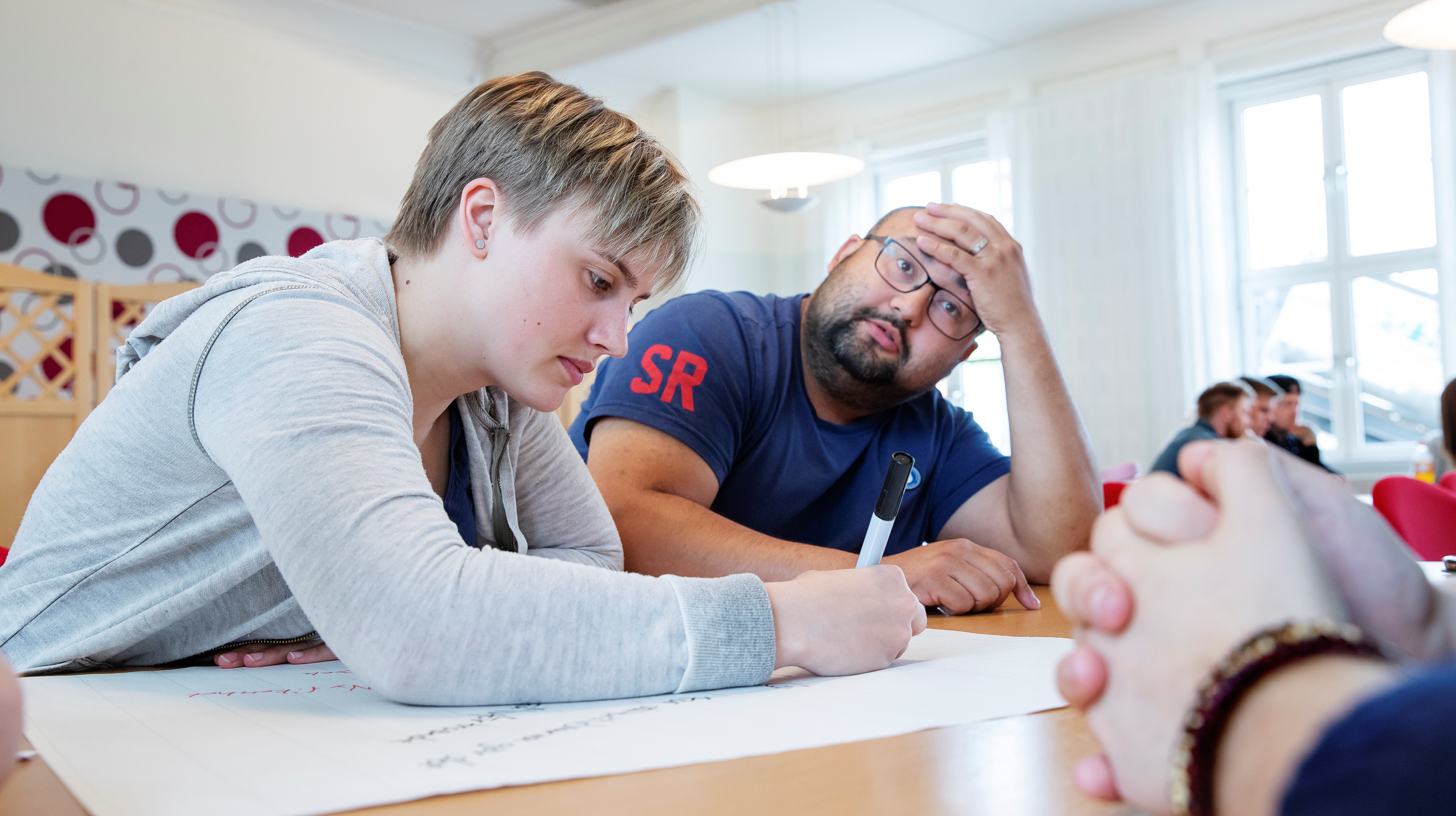 Två folkhögskoledeltagare i ett klassrum. Den ena ser ut att tänka högt och den andra noterar tankarna på ett papper.