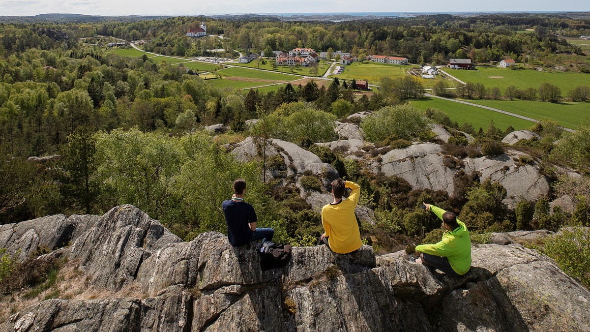 Tre folkhögskoledeltagare som sitter på en klippa och ser ut över ett landskap med både skog och fält. Billströmska folkhögskolan syns från utsiktsplatsen. 
